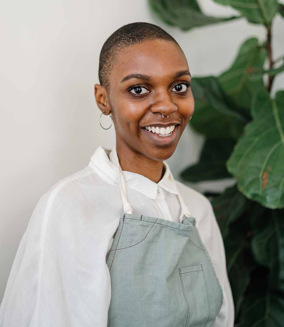 Industries African American Woman in an apron smiling into camera with plants behind her